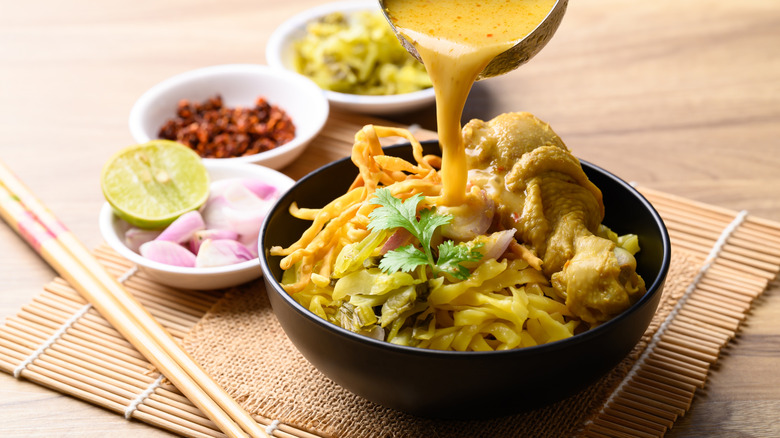 Ladling curry broth into a khao soi bowl, next to bowls filled with aromatics and spices