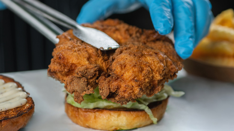 A gloved hand and set of tongs being used to place a fried chicken fillet onto a bun with lettuce