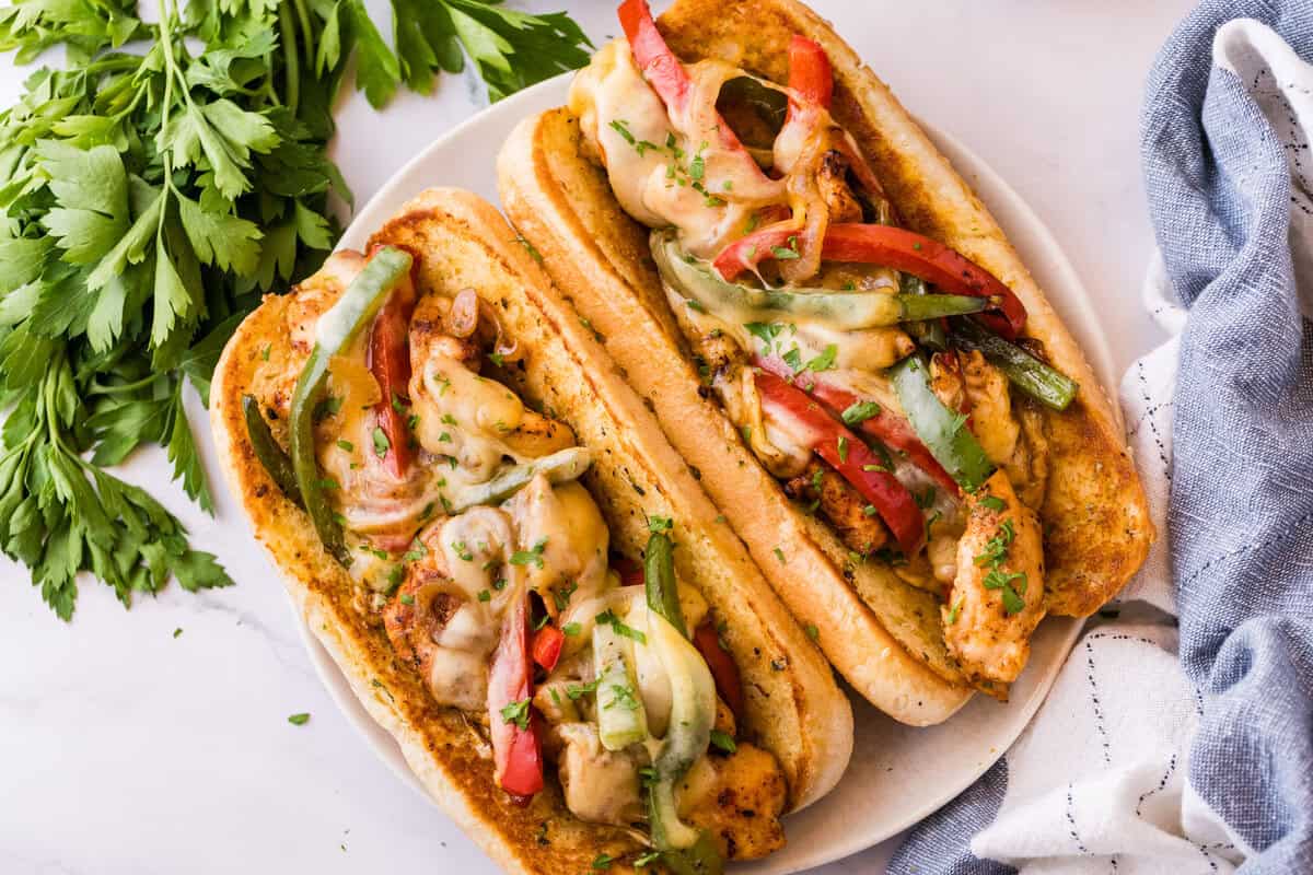 Udsyn fra oven af to kylling-cheesesteaks på en hvidt tallerken, med kylling, peberfrugt og smeltet provolone, pyntet med frisk persille og en blå stofhåndklæde ved siden af.