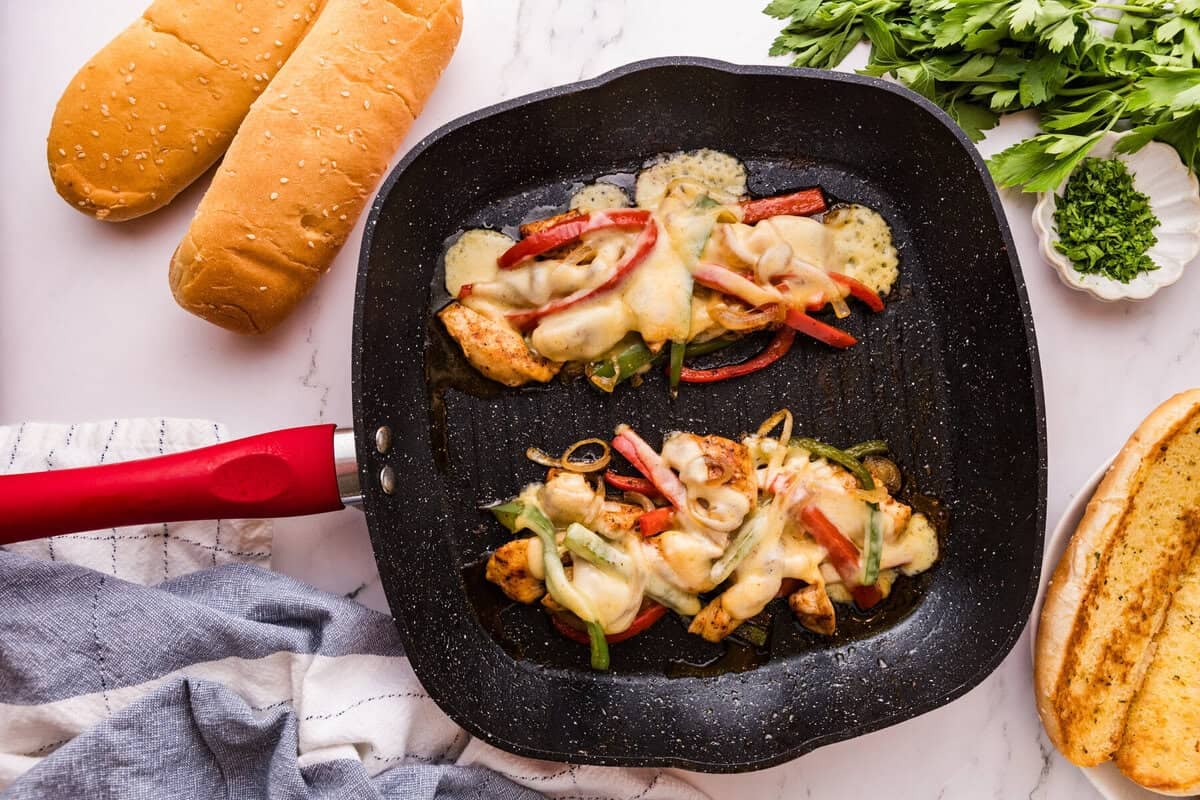 Provln længere smeltet over to portioner af kylling og peberfrugter i en grillpande, med hoagie-ruller, frisk persille og ristede hvidløgsbrød i nærheden.