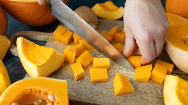 a person cubing butternut squash on a wooden cutting board surrounded by winter squashes