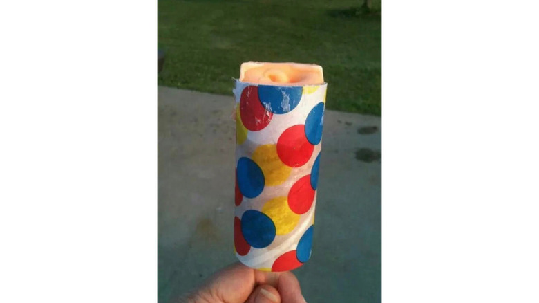A hand holding orange push up pop in polka dot rolled packaging against a driveway background