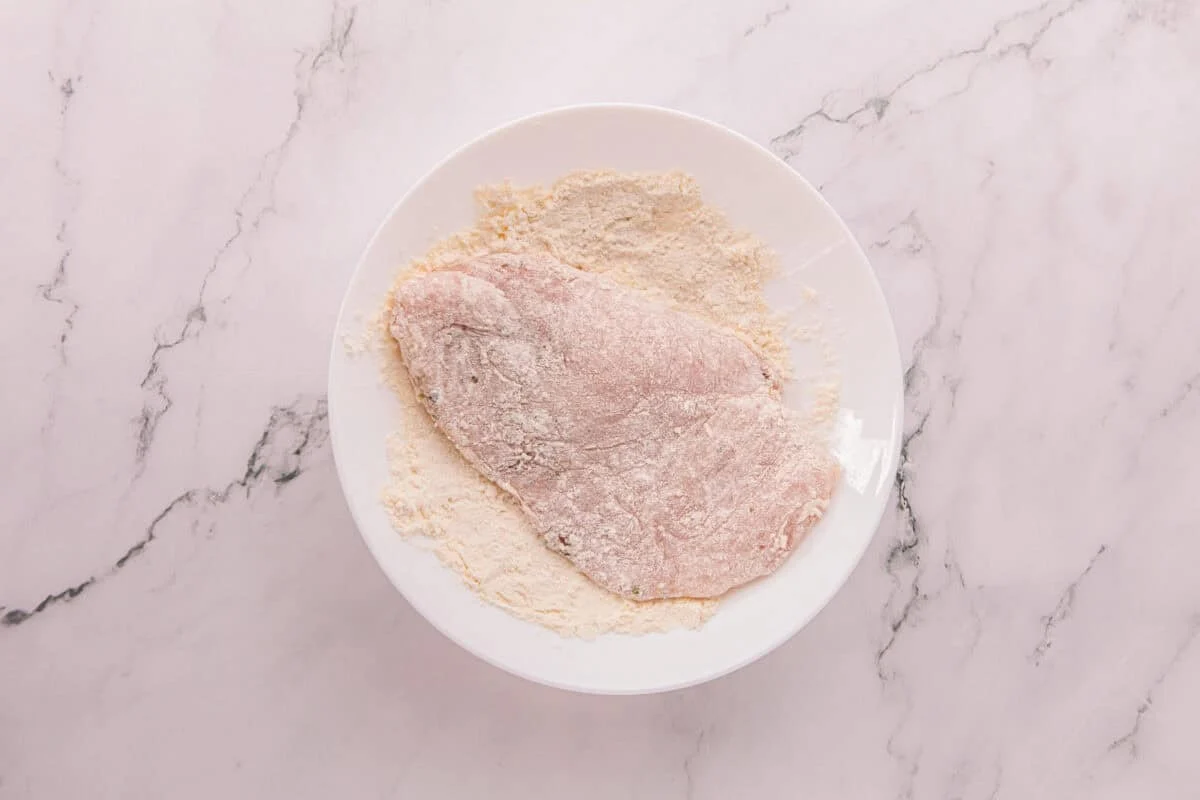A chicken breast being dredged in a flour and Parmesan mixture in a white bowl.