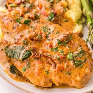 Close-up of creamy Tuscan chicken on a plate, topped with Parmesan cream sauce, sun-dried tomatoes, wilted spinach, and chopped parsley, served with mashed potatoes and asparagus.
