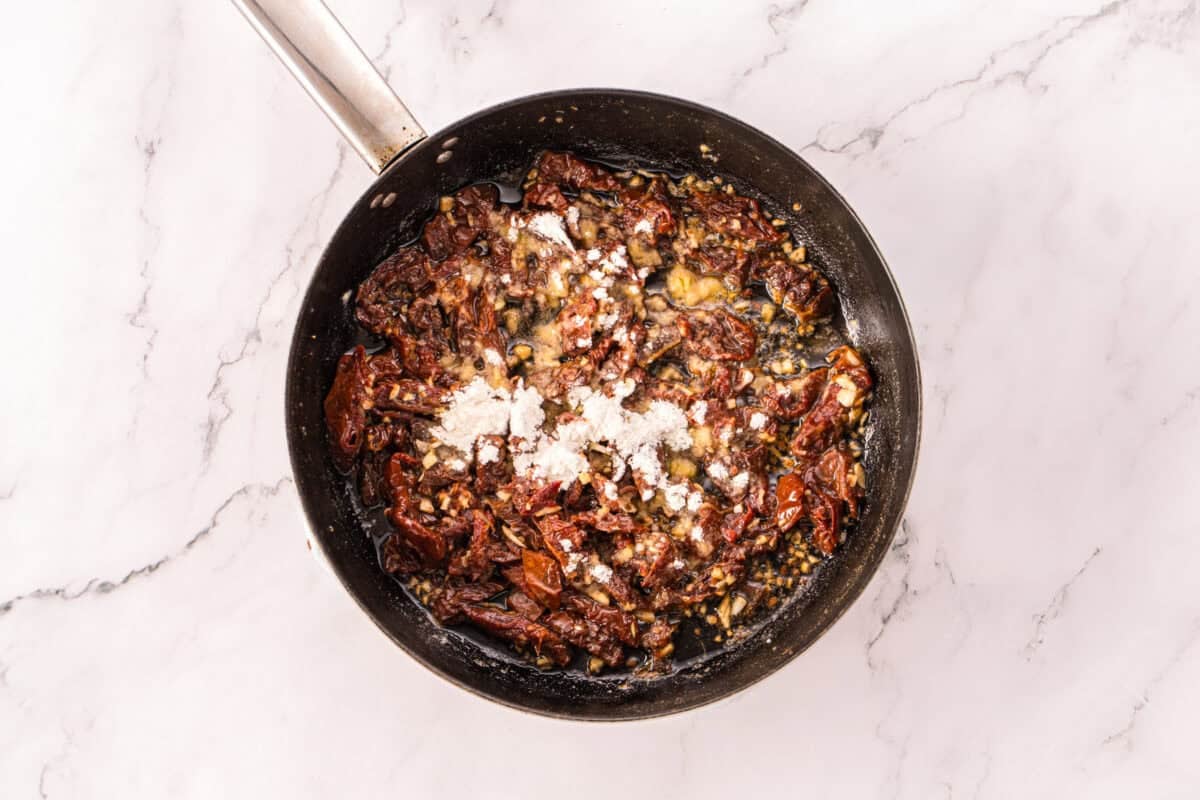 Flour sprinkled over sautéed sun-dried tomatoes and garlic in a skillet.