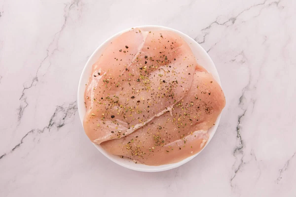 Raw chicken breasts in a white bowl, seasoned with Italian seasoning, salt, and black pepper.