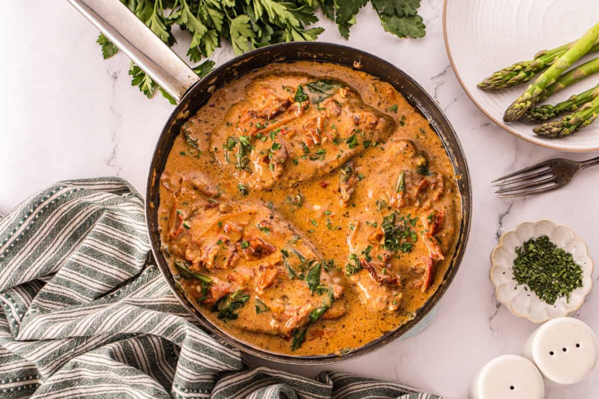 Overhead view of creamy Tuscan chicken in a skillet with sun-dried tomatoes, spinach, and parsley, next to a plate of asparagus and a striped kitchen towel.