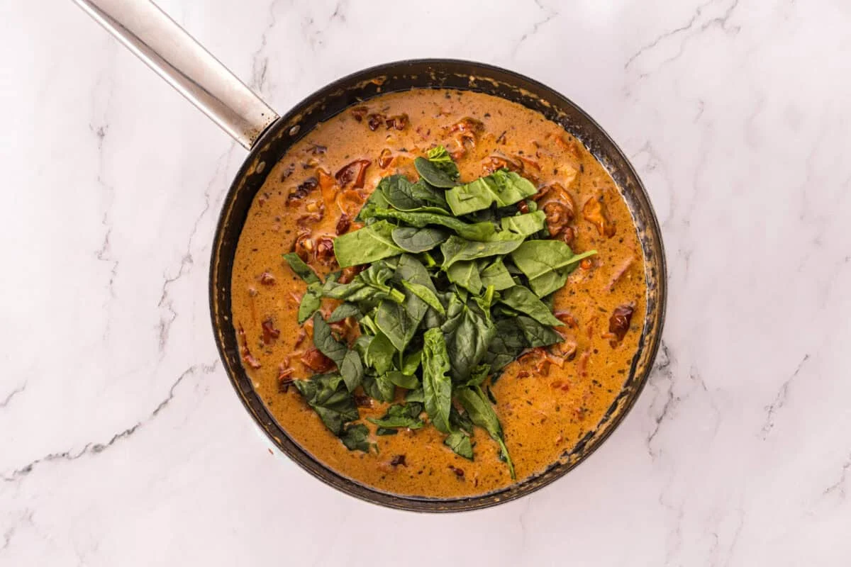 Fresh baby spinach piled on top of a creamy sun-dried tomato Parmesan sauce in a skillet.