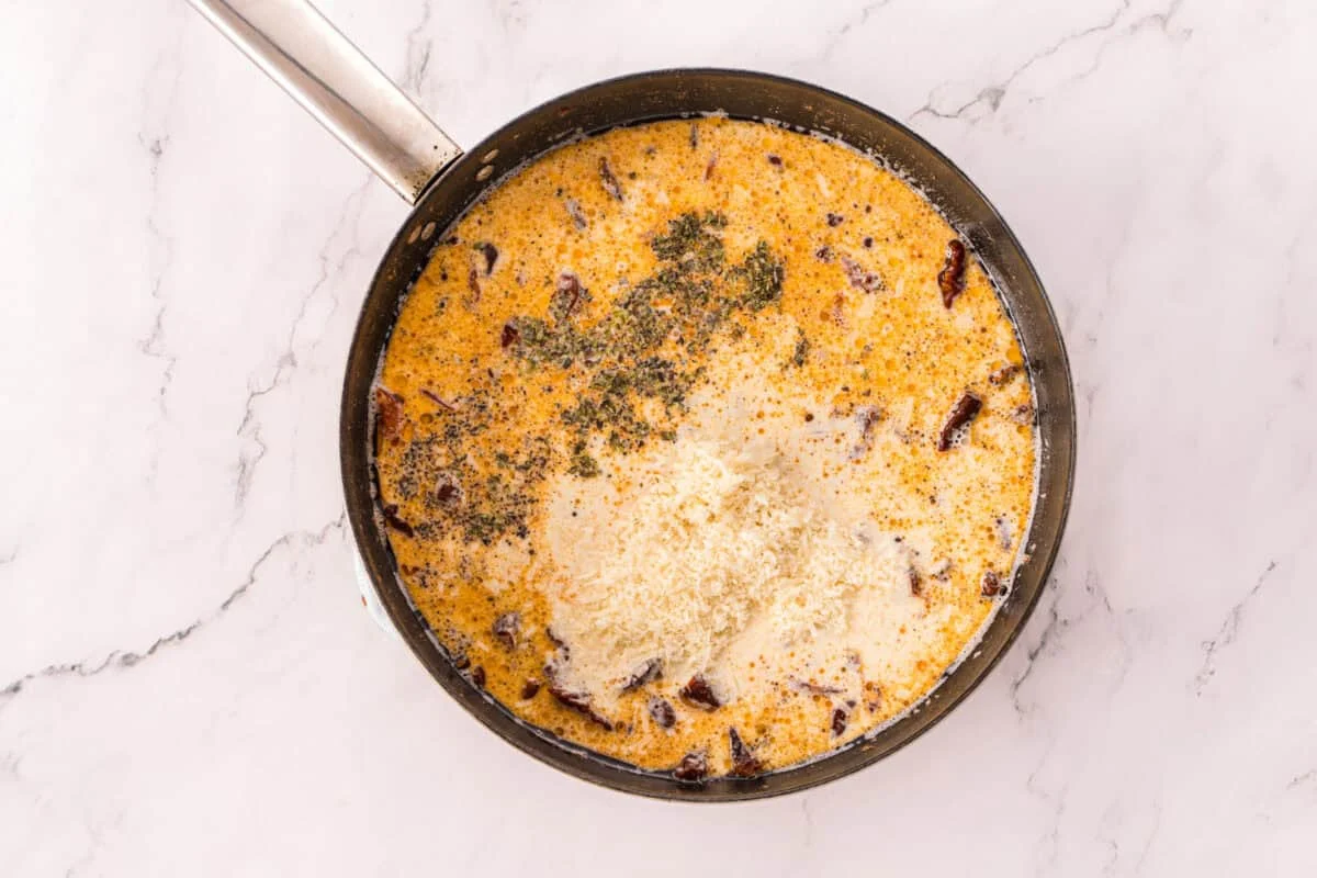 Heavy cream, chicken broth, grated Parmesan, Italian seasoning, and black pepper added to a skillet of sun-dried tomatoes before stirring.