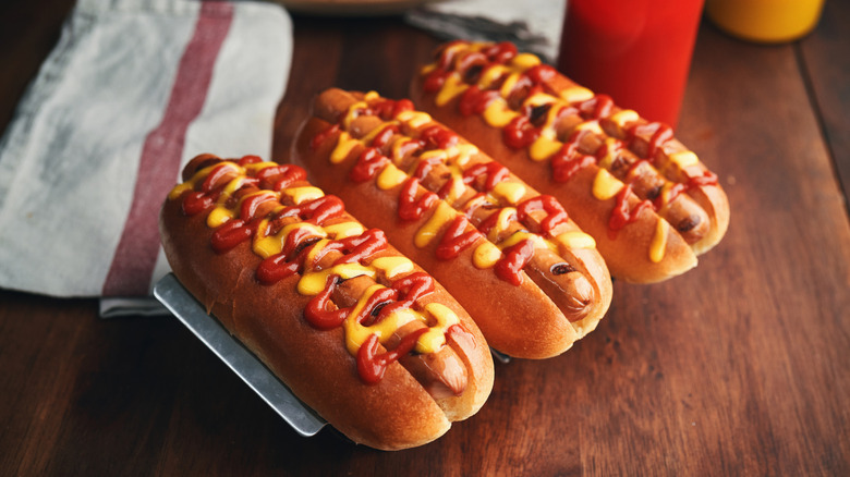 Three hot dogs drizzled with ketchup and mustard lined up on wooden table
