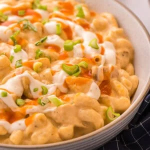 Side view of a bowl of buffalo chicken mac and cheese topped with ranch, buffalo sauce, and green onions next to a navy checkered towel.