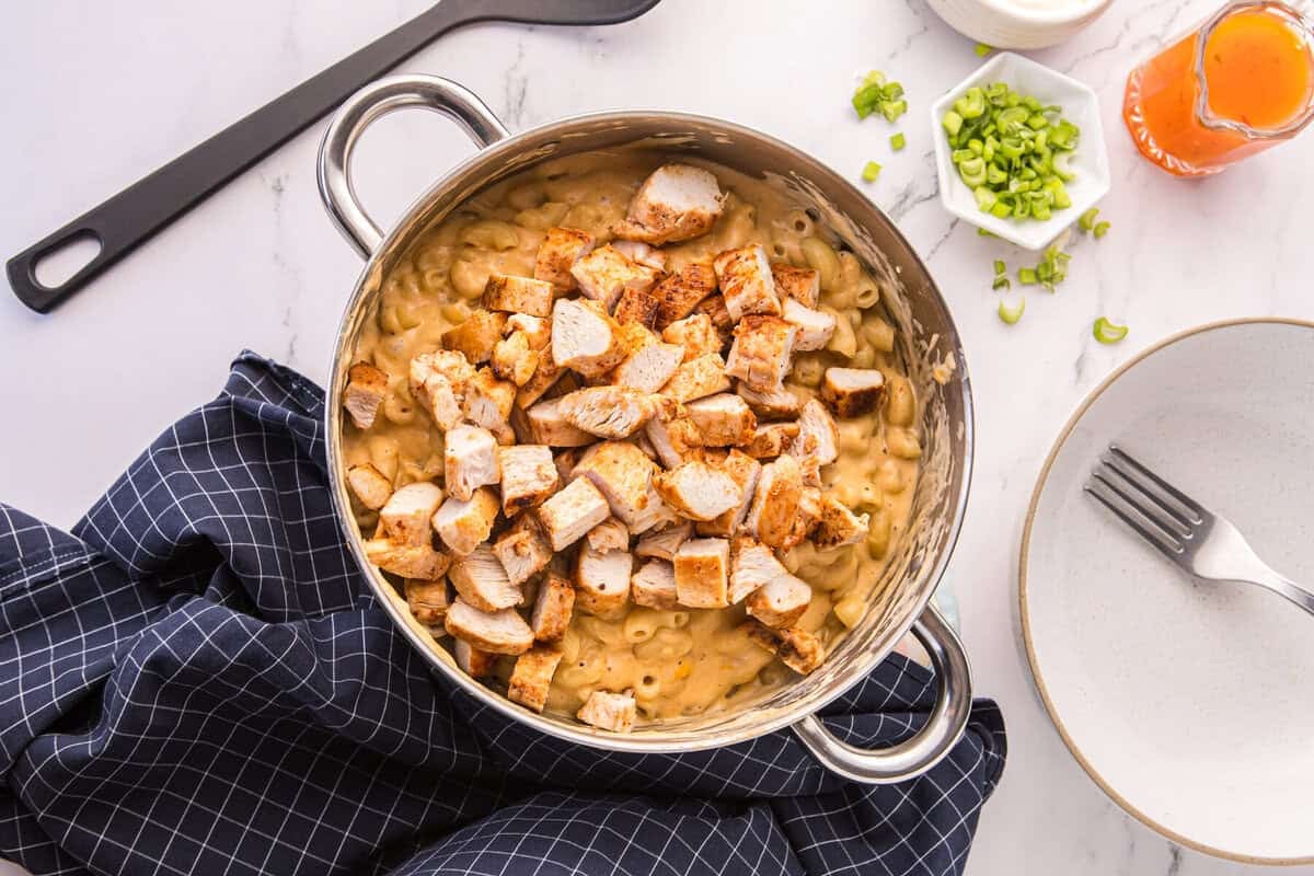 Chopped seared chicken added to a pot of creamy buffalo mac and cheese, with green onions and buffalo sauce on the side.