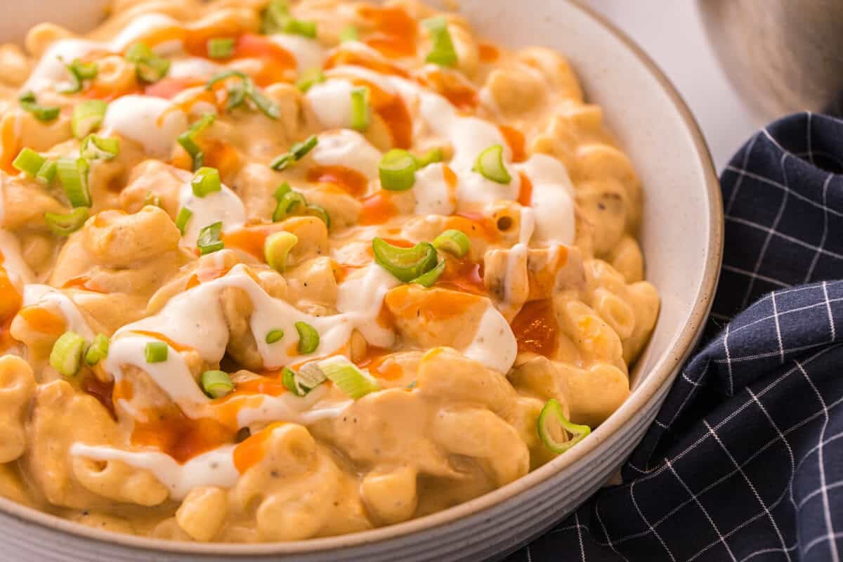 Side view of a bowl of buffalo chicken mac and cheese topped with ranch, buffalo sauce, and green onions next to a navy checkered towel.