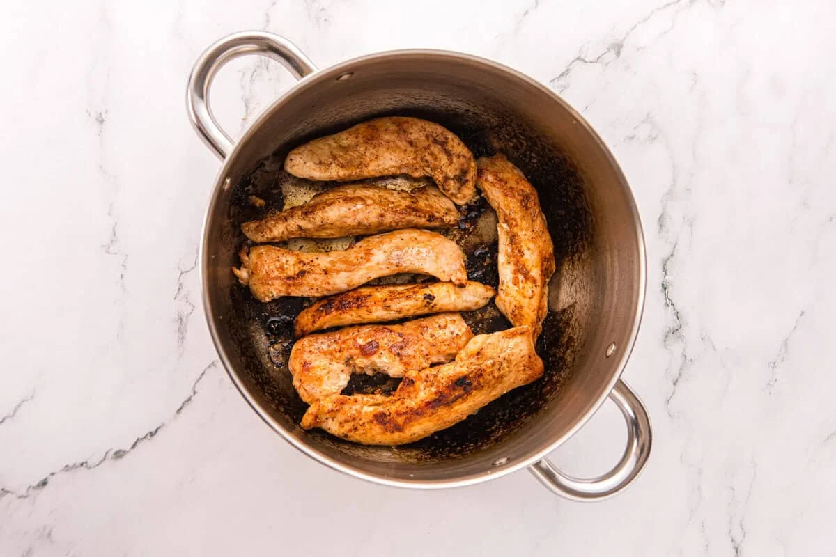 Seasoned chicken tenders seared golden brown in a stainless steel pot with browned bits on the bottom.
