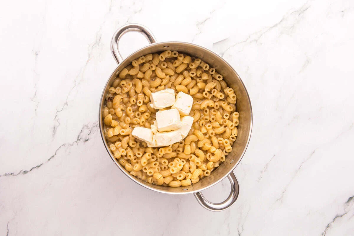 Cubes of cream cheese on top of cooked elbow macaroni in a stainless steel pot.