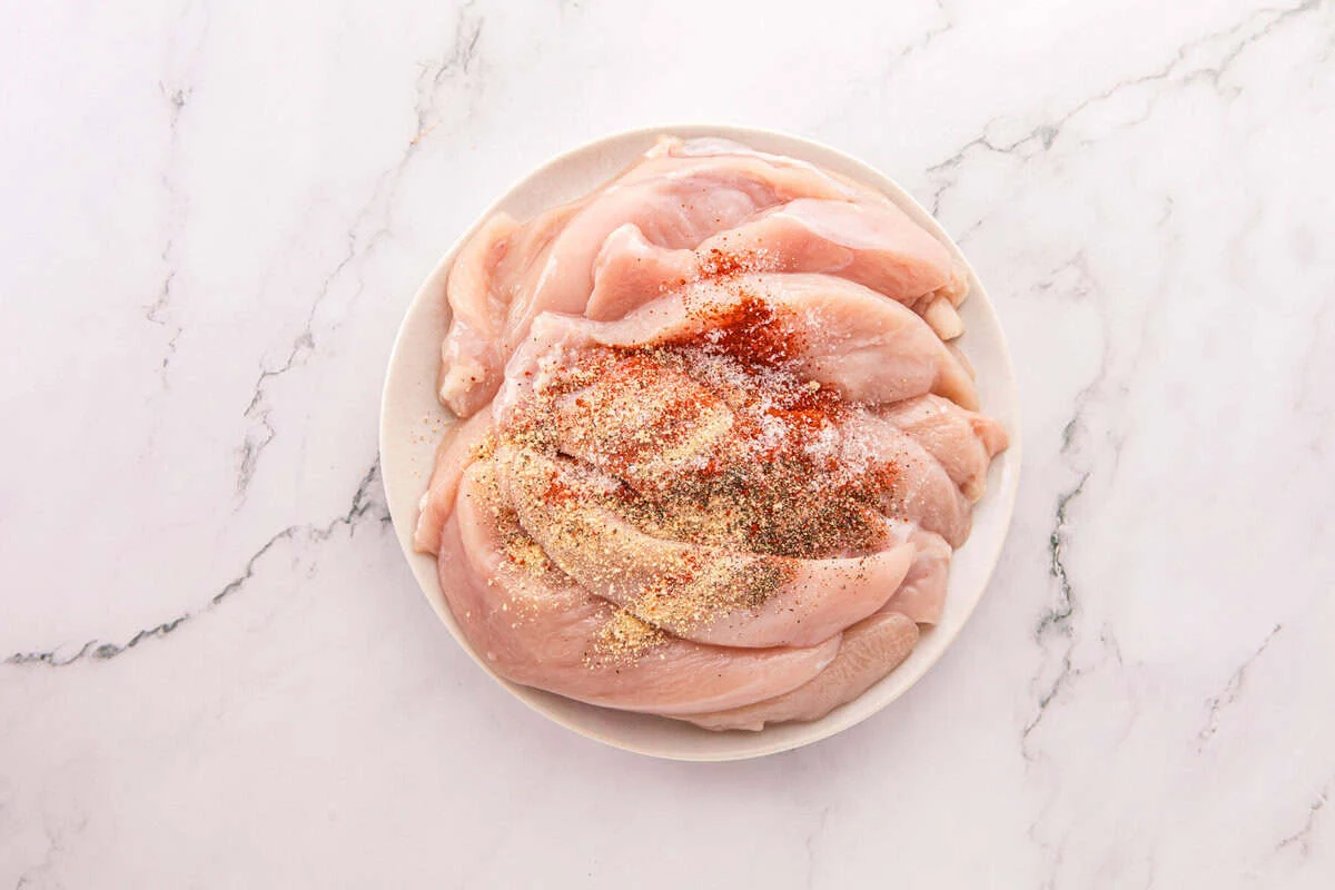 Raw chicken tenders on a white plate sprinkled with garlic powder, paprika, salt, and black pepper.