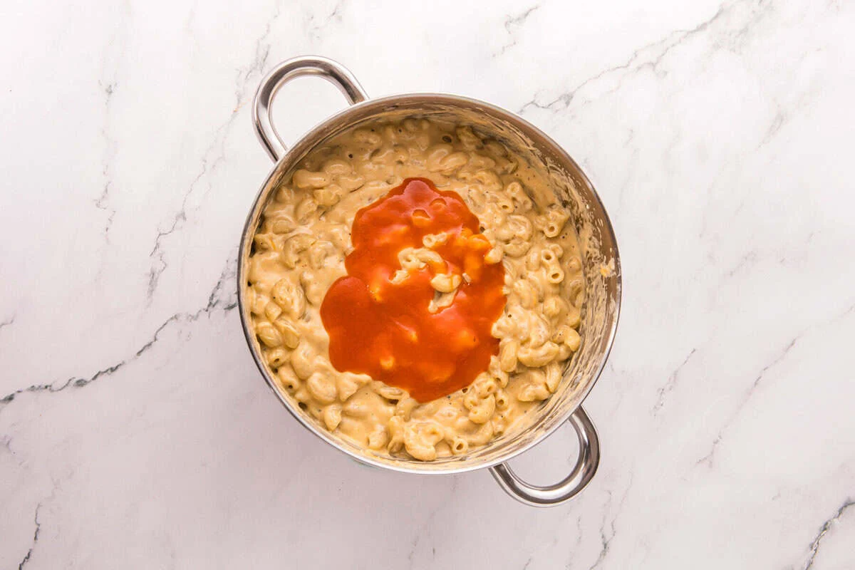 Buffalo sauce poured over creamy mac and cheese in a stainless steel pot.