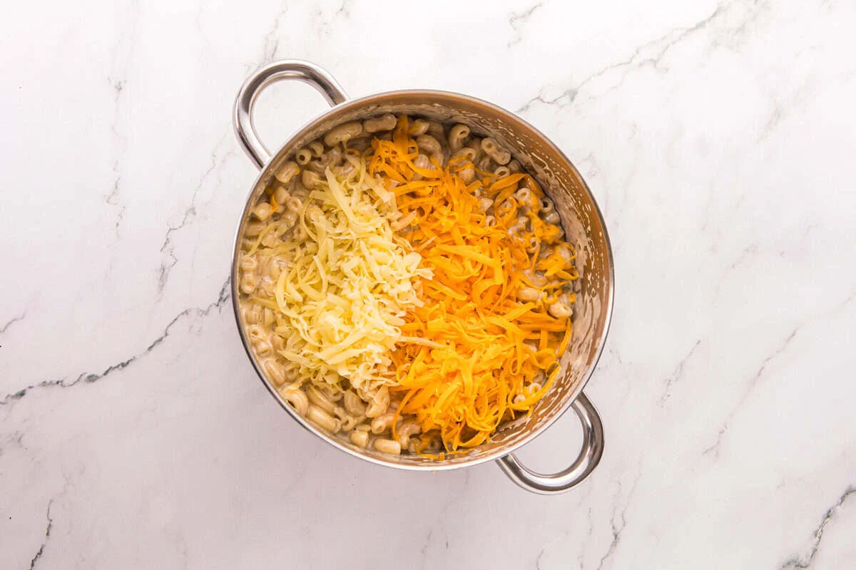 Freshly shredded sharp cheddar and Monterey Jack cheese piled on top of creamy macaroni in a pot.