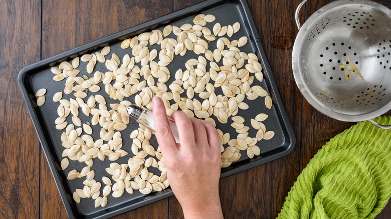 Hand sprinkling salt onto pumpkin seeds on baking sheet next to a colander