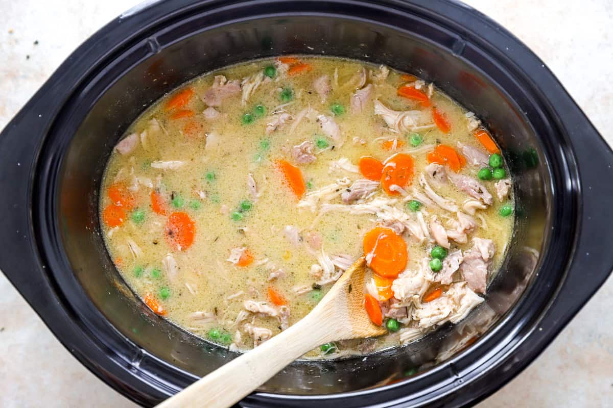 Shredded chicken, carrots, and peas stirred into a creamy broth in a slow cooker with a wooden spoon.