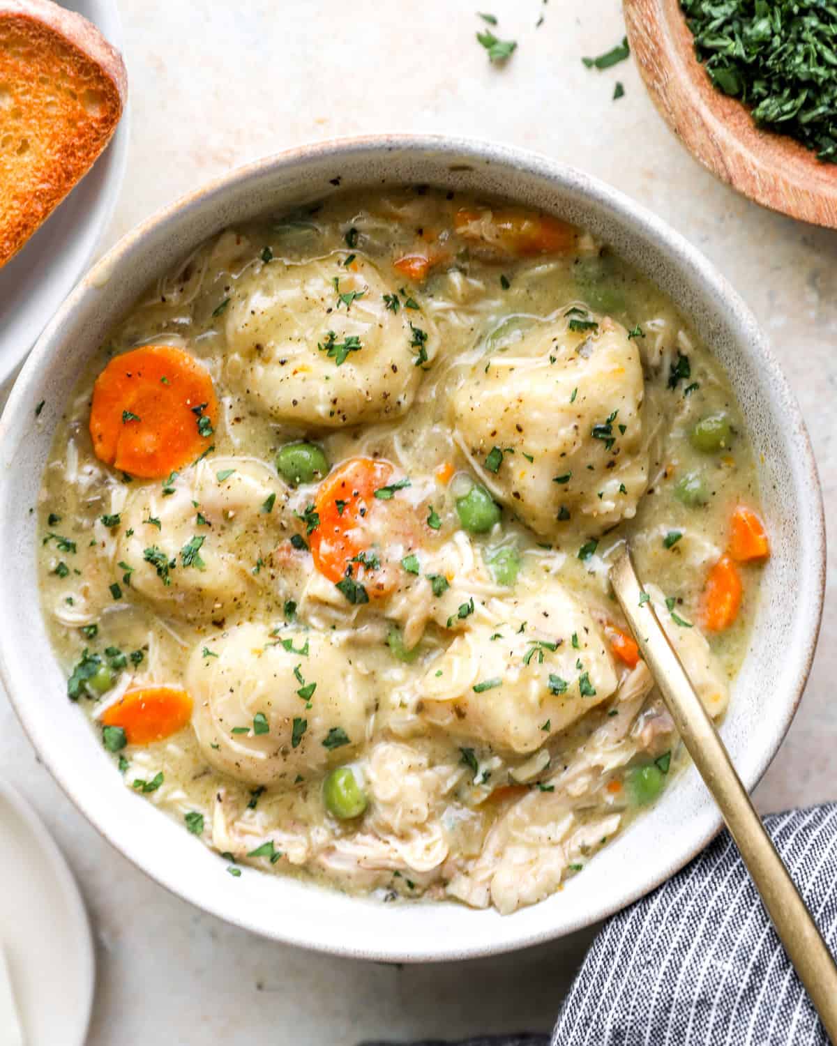 Overhead bowl of crockpot chicken and dumplings with fluffy biscuit dumplings, carrots, peas, and fresh parsley beside toasted bread.
