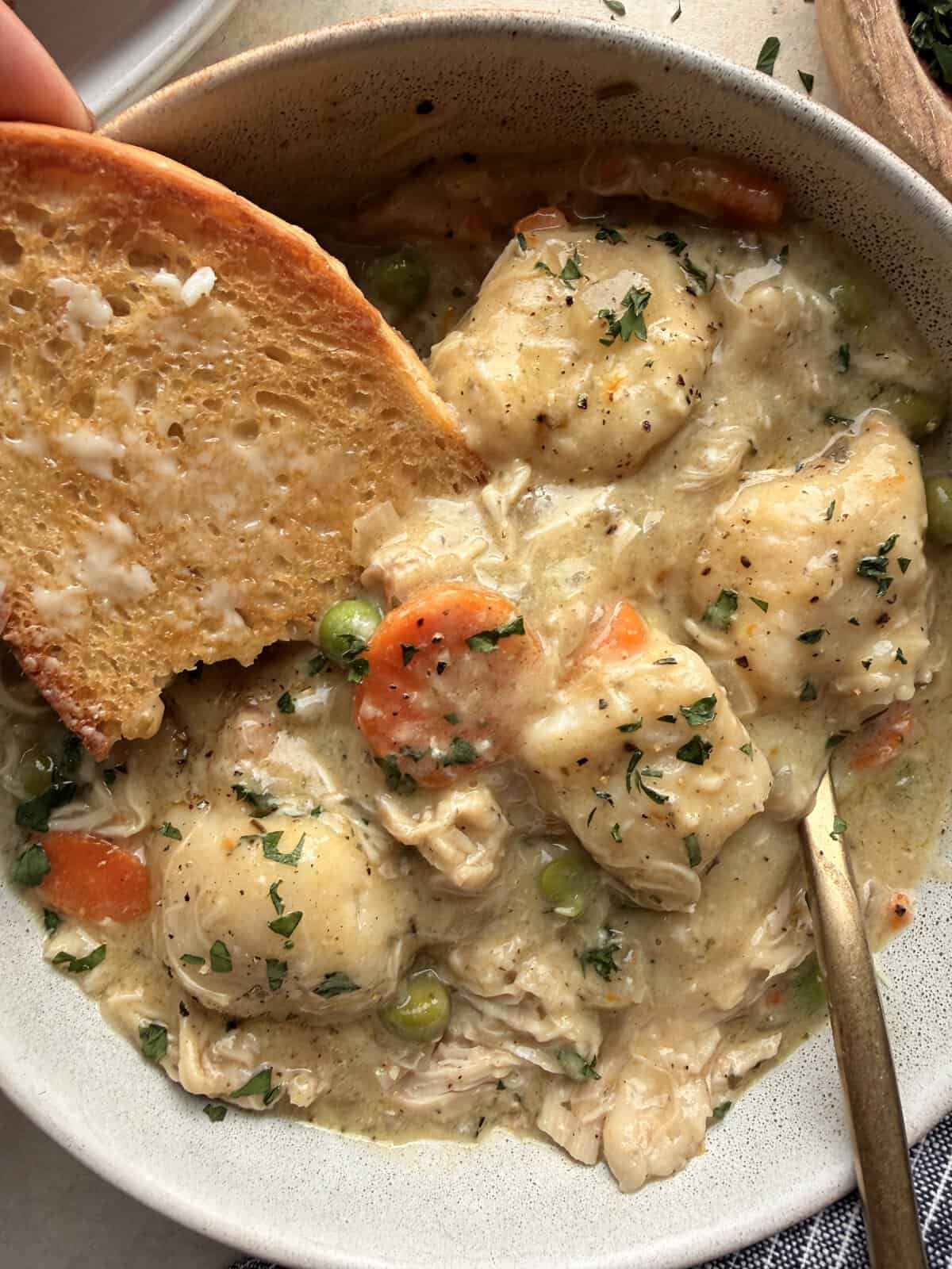 Buttered toasted bread dipped into a bowl of creamy chicken and dumplings with carrots and peas.