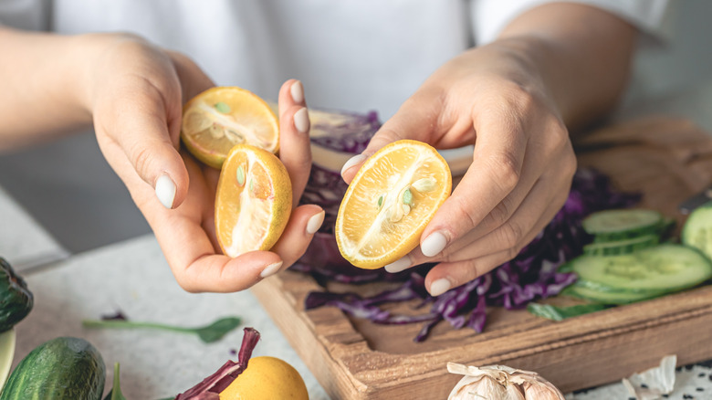 A person holds halved limequats over a cutting board with red cabbage and cucumbers
