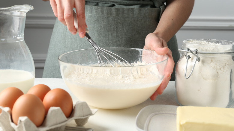 Hands whisking a bowl of cake mix with flour, butter, eggs, and milk nearby