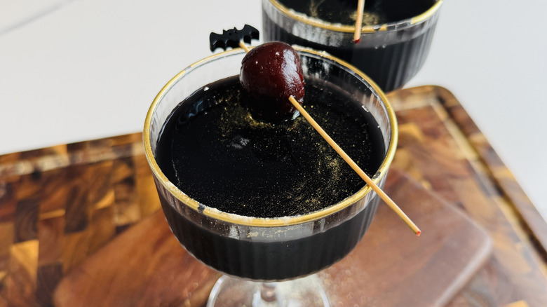 Black magic cocktail in glass garnished with cherry and gold luster dust