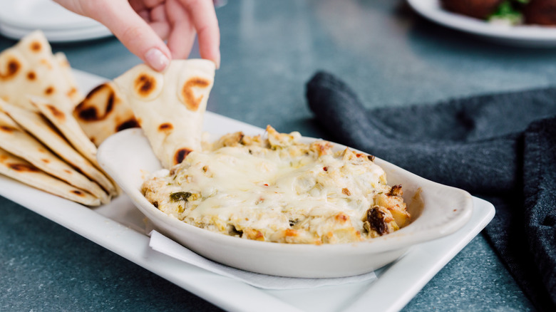 Someone dipping a pita wedge into a plate of artichoke dip