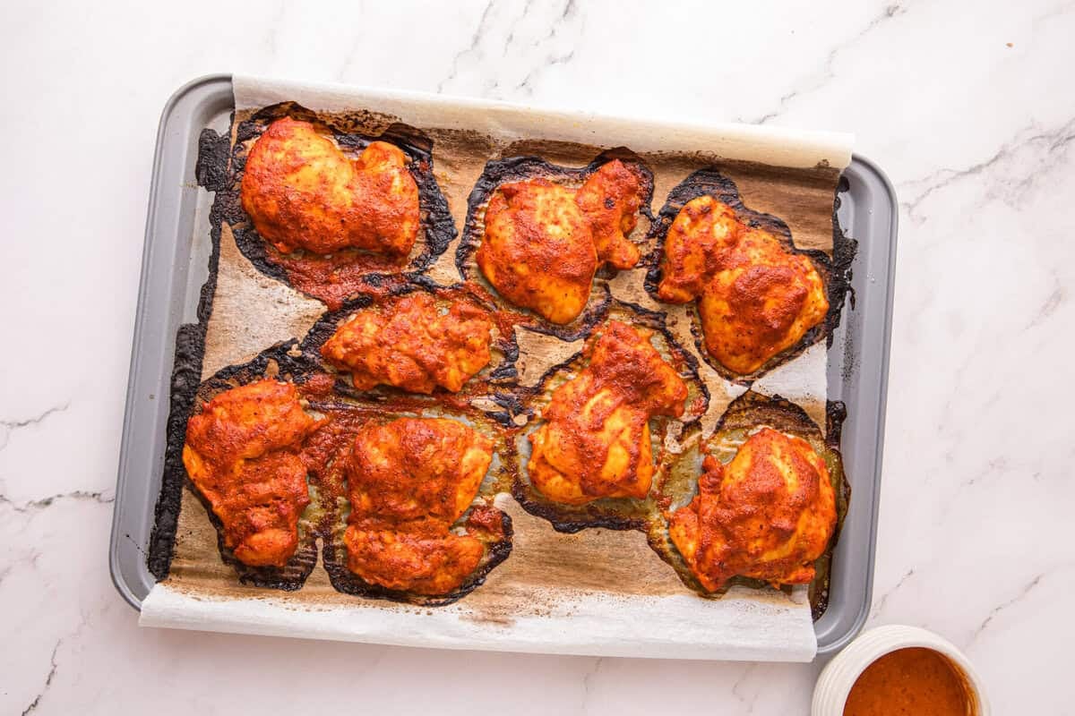 Baked peri peri chicken thighs with charred edges on a sheet pan, with finishing sauce in a bowl beside it.