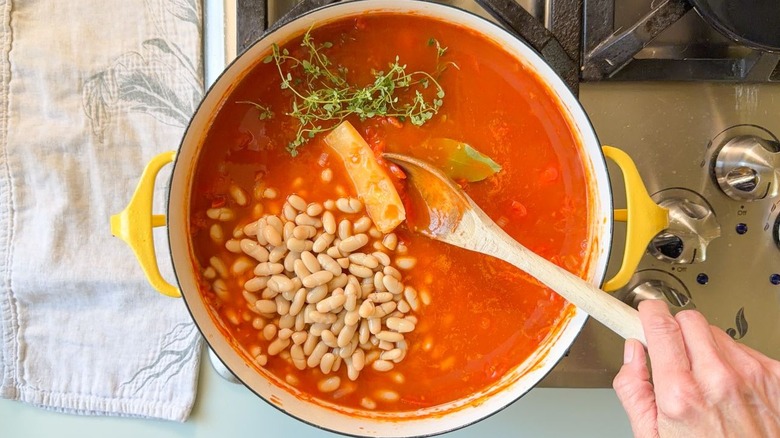 Adding canned white beans, thyme sprigs, bay leaf, and a parmesan rind to tomato soup ingredients in Dutch oven on stovetop