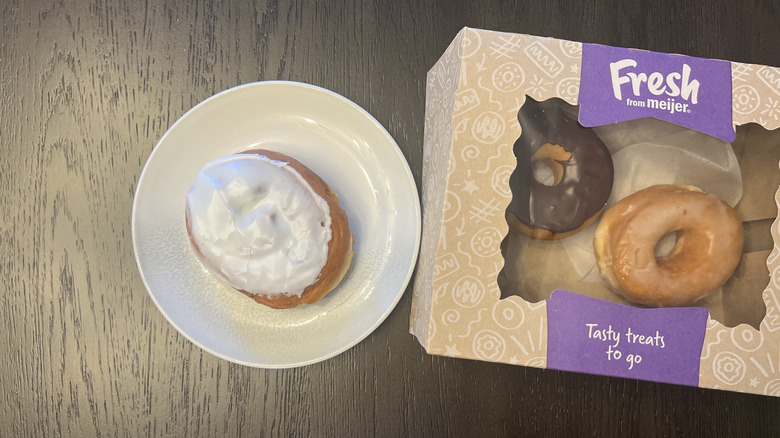 A box of Meijer donuts on a wooden table, next to a white plate with a white iced cinnamon donut