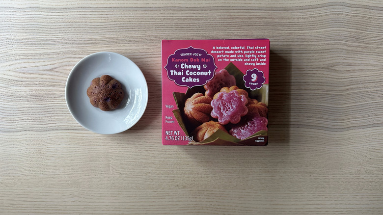 A box of Trader Joe's Chewy Thai Coconut Cakes on a wooden table, next to a single cake on a white plate to the left of the box