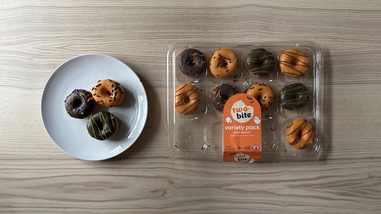 A box of mini donuts frosted with orange and chocolate icing and dusted with sprinkles on a wooden table, next to three donuts on a white plate