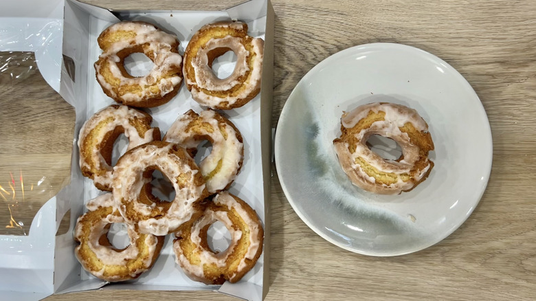 A box of Walmart's Glazed Sour Cream Donuts open on a wooden table, next to a single donut on a white plate