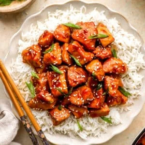 Overhead view of bourbon chicken over white rice on a scalloped plate, topped with sesame seeds and green onions, with chopsticks.