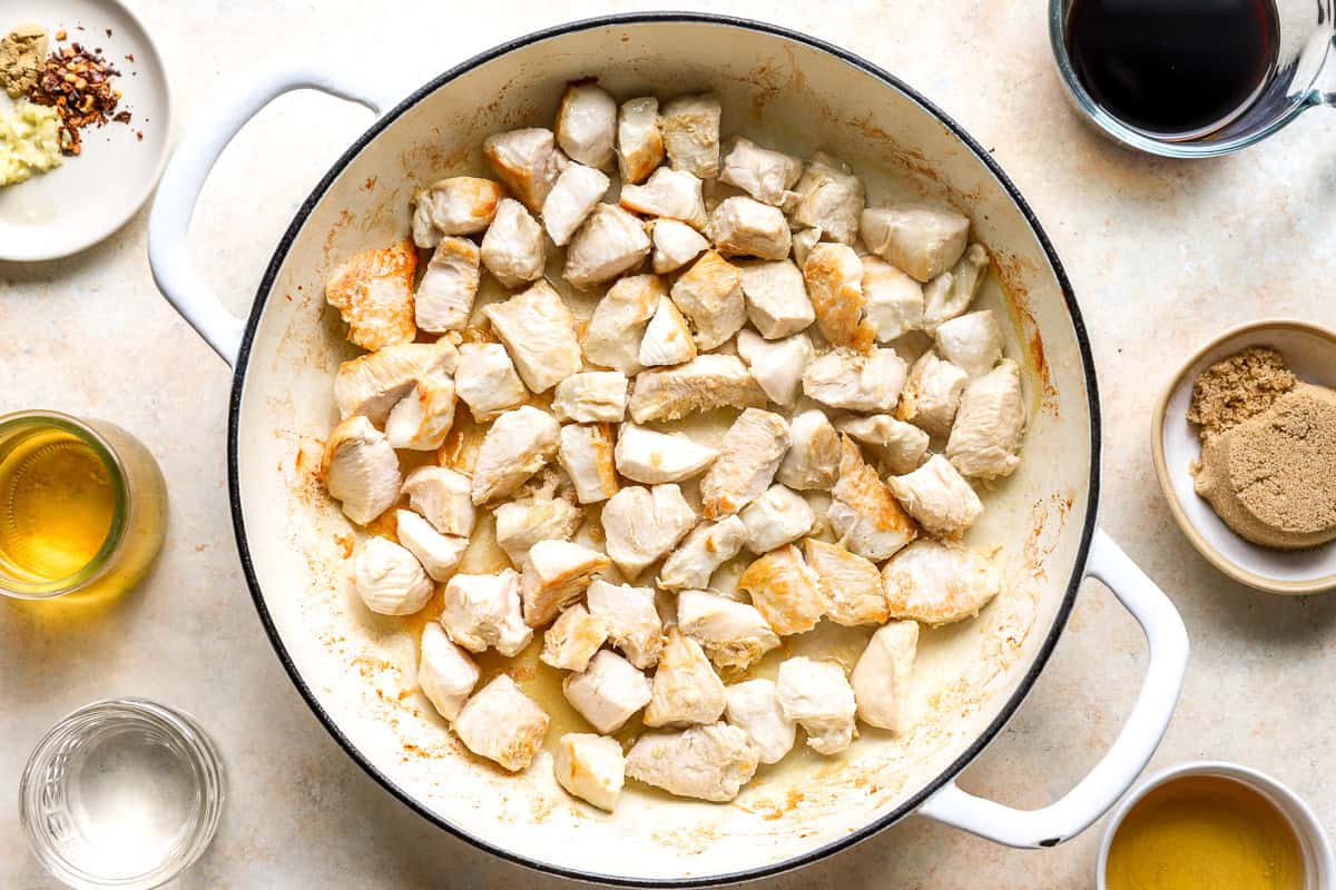 Lightly browned chicken breast pieces in a white skillet, surrounded by bowls of brown sugar, soy sauce, garlic, and red pepper flakes.