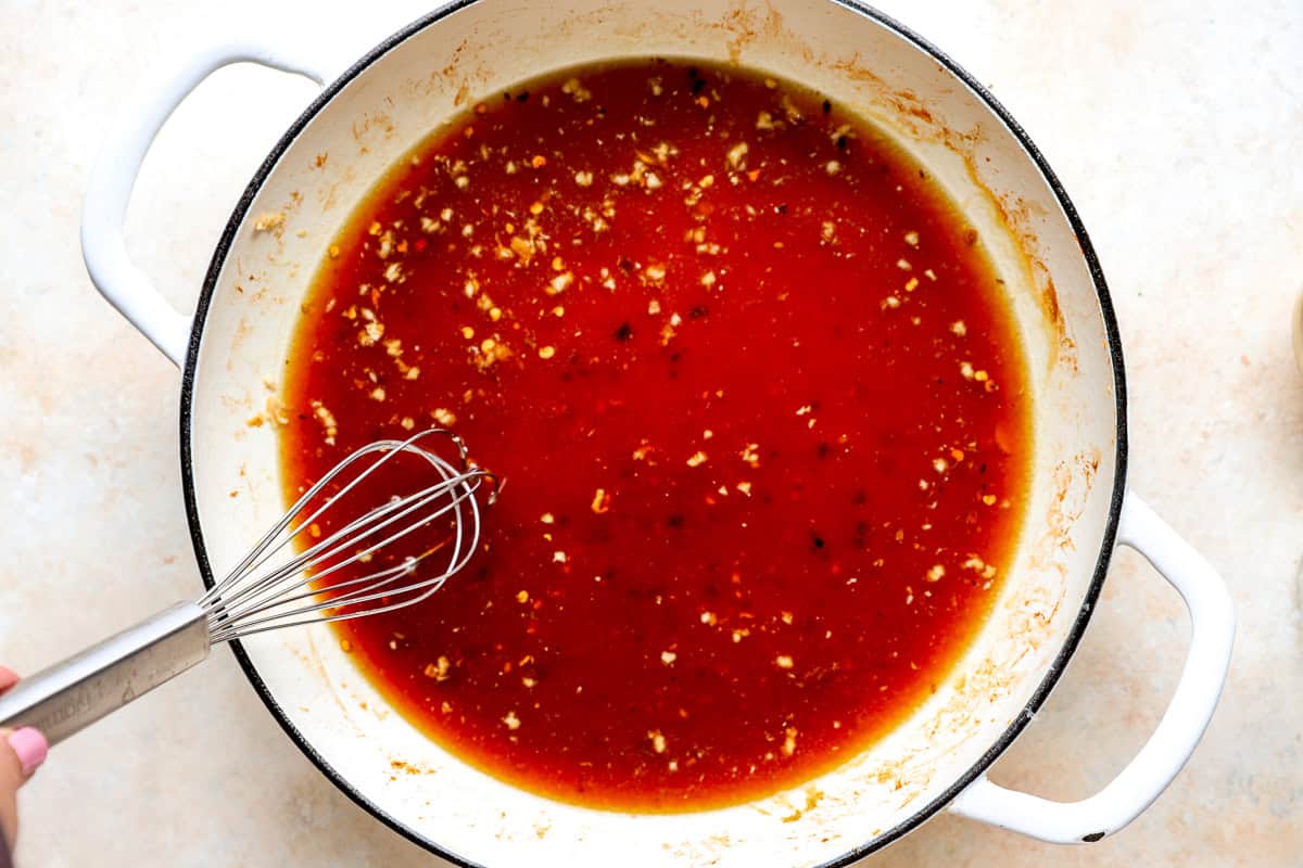 A whisk stirring bourbon sauce with garlic and red pepper flakes in a white skillet.
