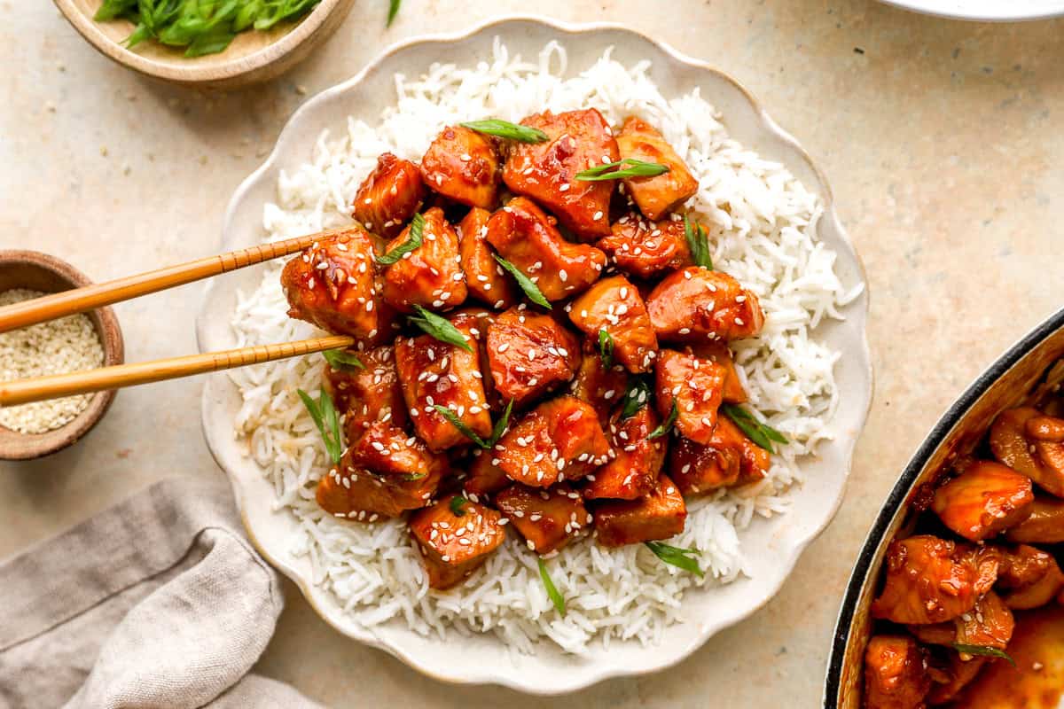 Chopsticks lifting a piece of glazed bourbon chicken from a plate of white rice topped with sesame seeds and green onions.