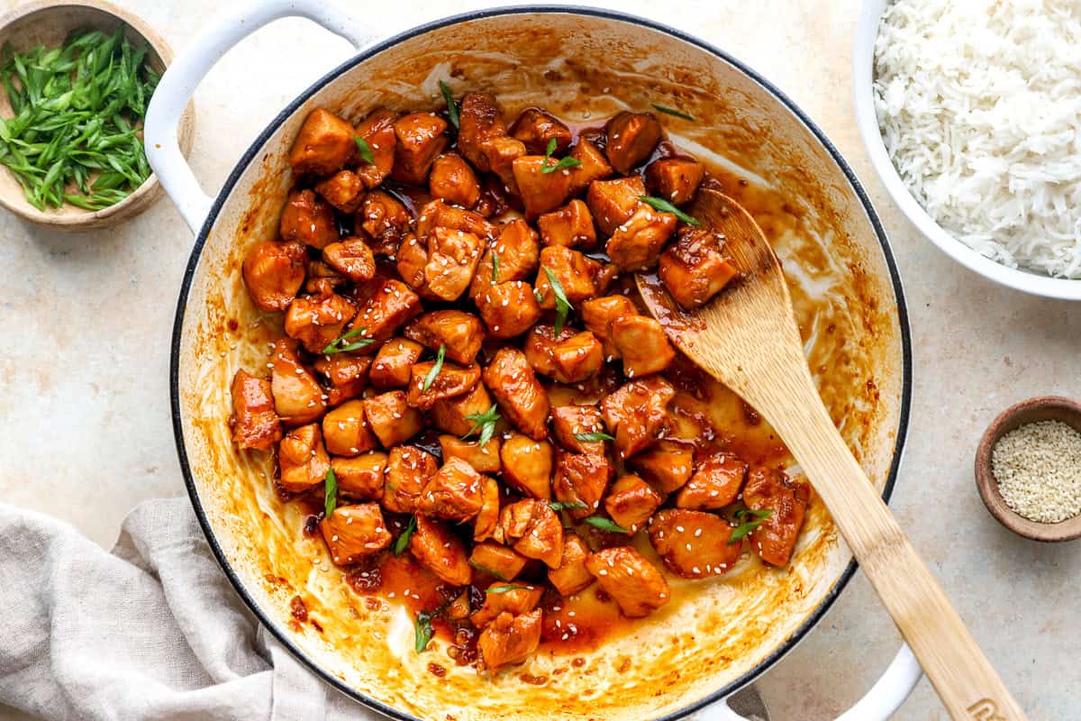 Glossy bourbon chicken in a skillet topped with sesame seeds and green onions, next to bowls of rice, sesame seeds, and green onions.