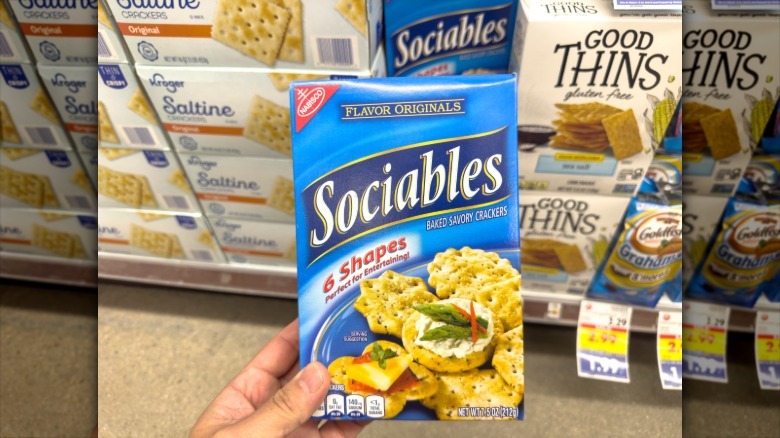 A hand holds a box of Sociables in a grocery store cracker aisle.