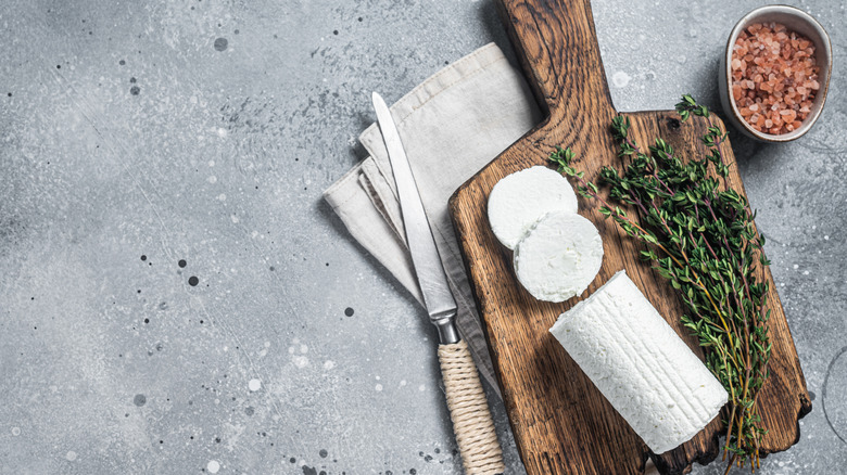 a log of goat cheese with two slices sits on a wooden cutting board with sprigs of thyme, a knife next to. it, and a container of salt on the other side.
