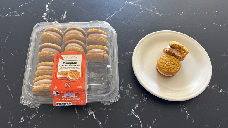 A Bake Shop Pumpkin Cookie Sandwiches container next to two cookie sandwiches, one bitten, on a white plate