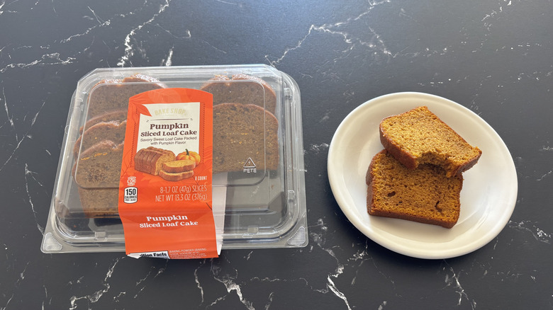 A container of Bake Shop Pumpkin Sliced Loaf Cake next to two loaf slices, one bitten, on a white plate