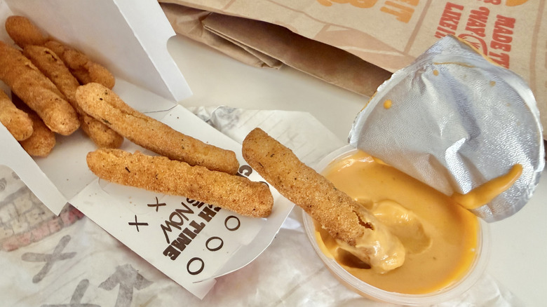 Chicken fries spill out of a box. One is being dipped into the nacho cheese sauce.