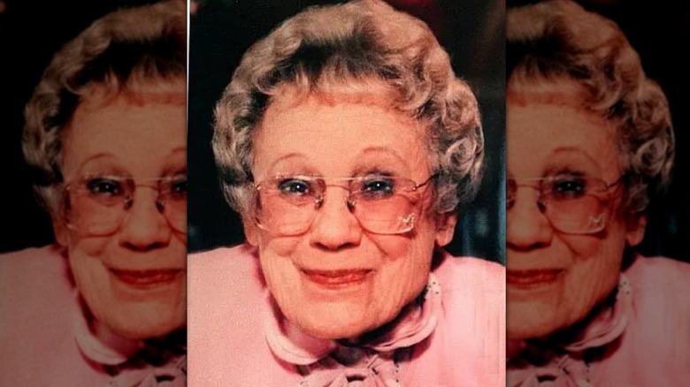 Older woman with short gray hair, red lipstick, and glasses, wearing a pink shirt and smiling