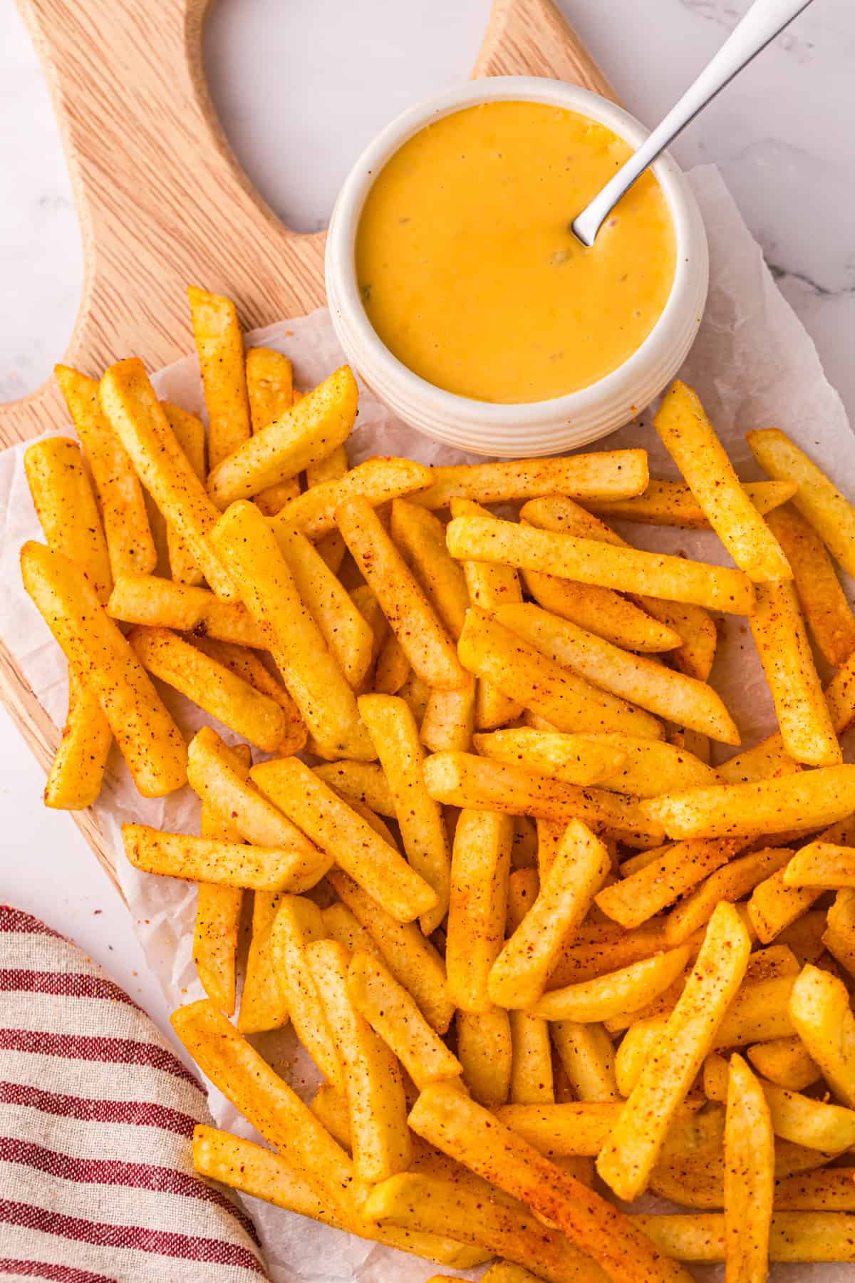 Seasoned nacho fries piled on a parchment-lined wooden board next to a small white bowl of creamy nacho cheese with a spoon, with a red striped kitchen towel at the edge.