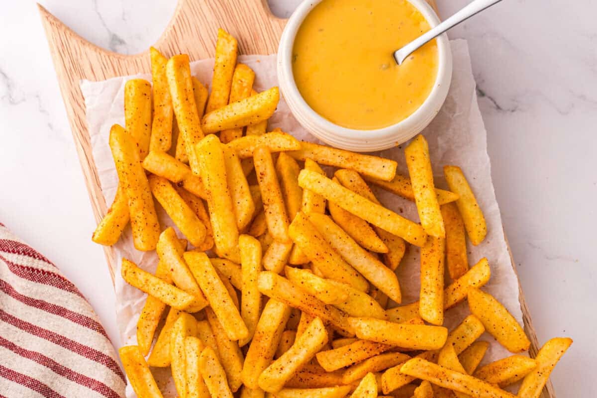 Crispy seasoned nacho fries spread across a wooden serving board with a bowl of warm nacho cheese for dipping.