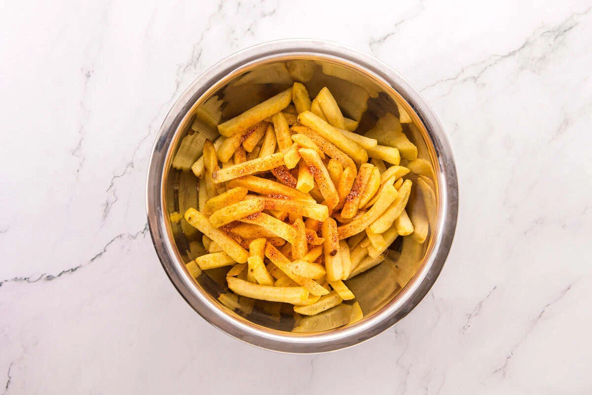 Freshly cooked fries in a stainless steel mixing bowl, sprinkled with reddish-orange nacho fry seasoning before tossing.
